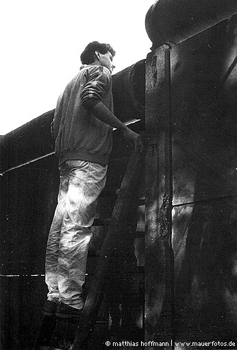 Mauerfoto: Ein Mann steht auf einer an die Mauer gelehnt Leiter und schaut auf die andere Seite.